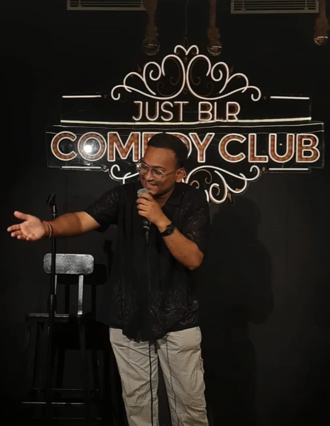Siddharth performing with a microphone in front of a Just BLR Comedy Club sign.