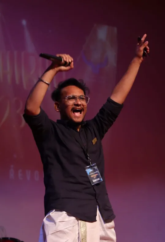 Siddharth on stage with both arms raised, holding a microphone.