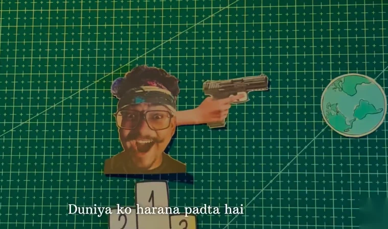 A paper cut-out collage on a green cutting mat: a cut-out of my head with an arm raised, holding a pistol, a paper globe at the right, and a paper podium numbered 1, 2 and 3 at the bottom, with the subtitle “Duniya ko harana padta hai” (“the world has to be beaten”).